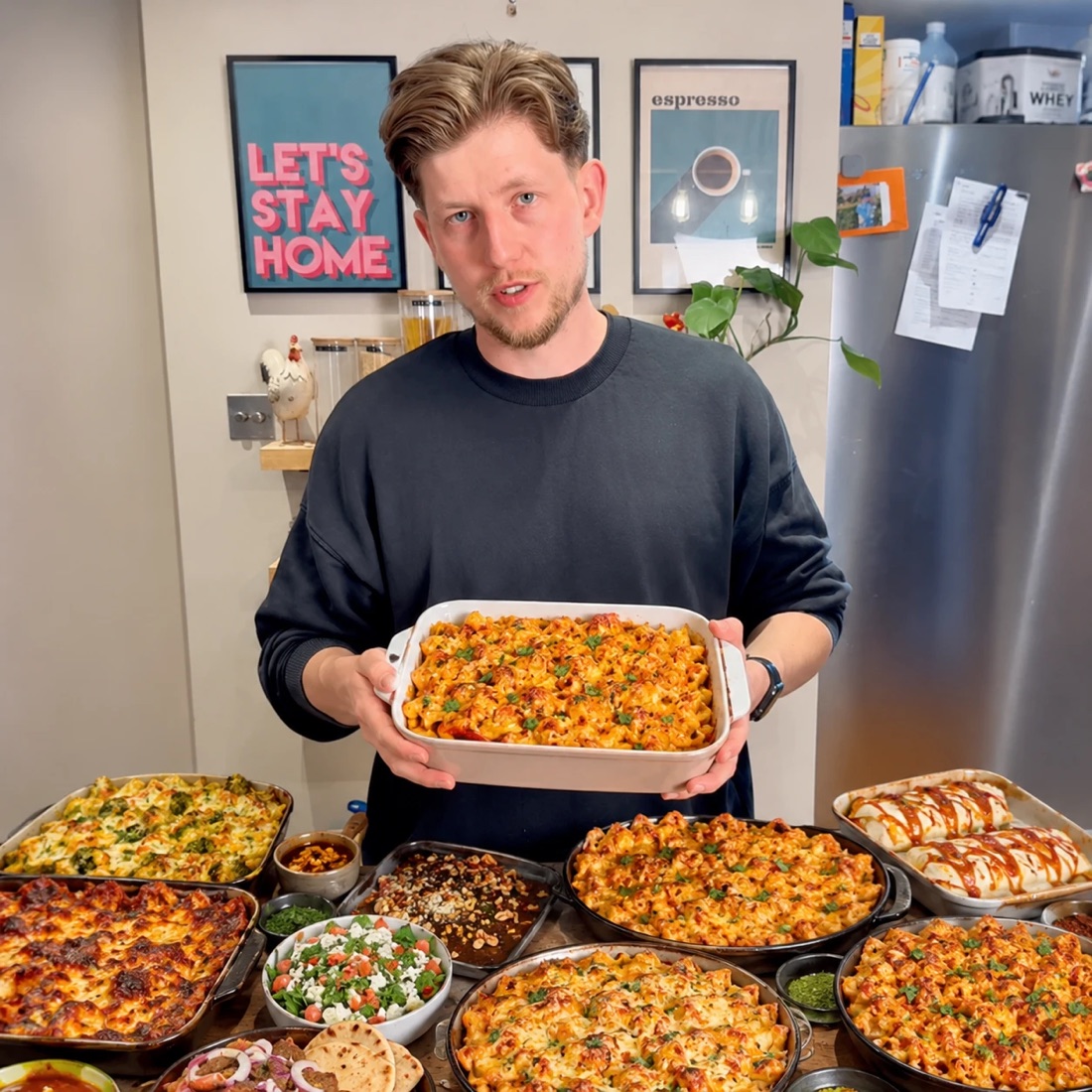 Jake in his kitchen holding a pasta bake, with a table full of family traybakes